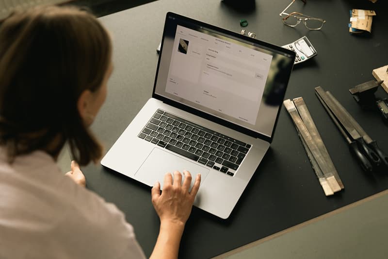 A person's hand operates a laptop on a dark desk surrounded by metal tools and glasses