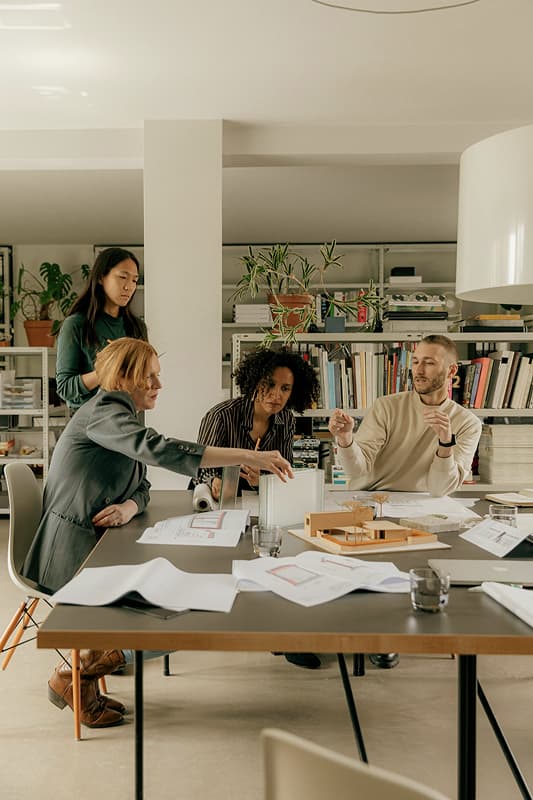 Four professionals discuss plans around a table with architectural models and documents